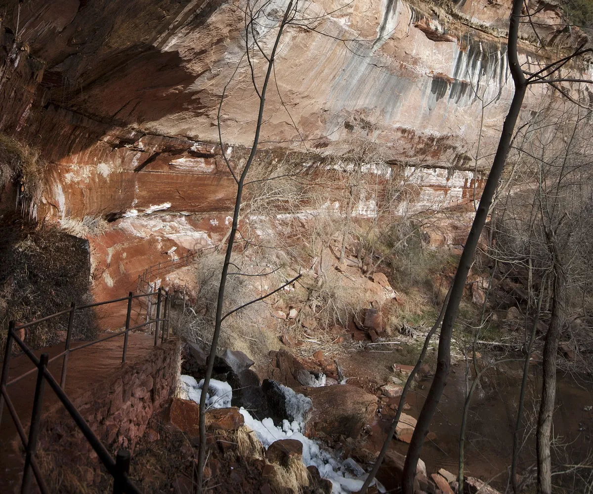 Lower Emerald Pool trail railing beneath a red sandstone overhang with ice and snow below