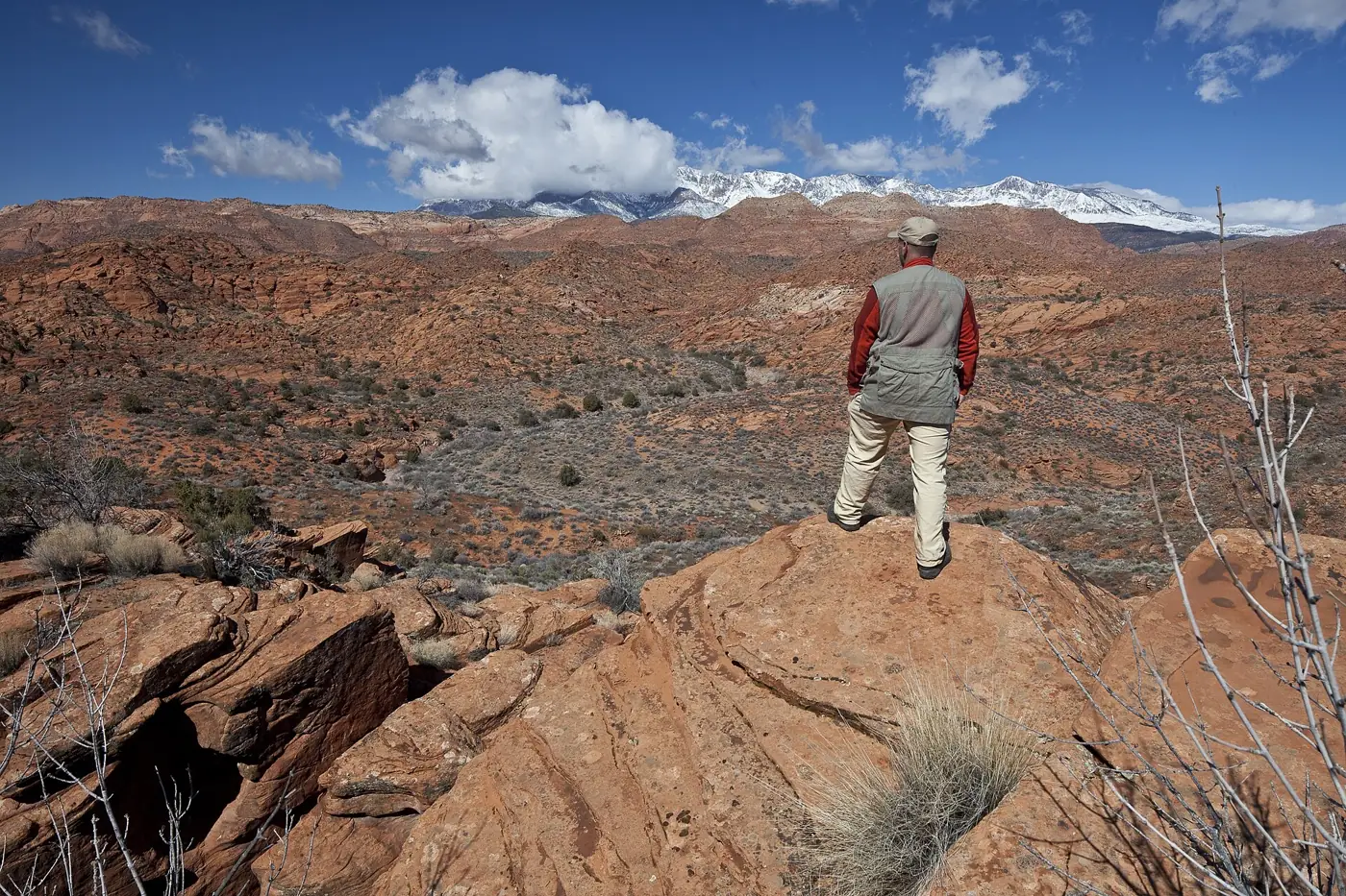 A hiker standing on red rock overlooking Red Cliffs National Conservation Area with snow-capped mountains in the distance