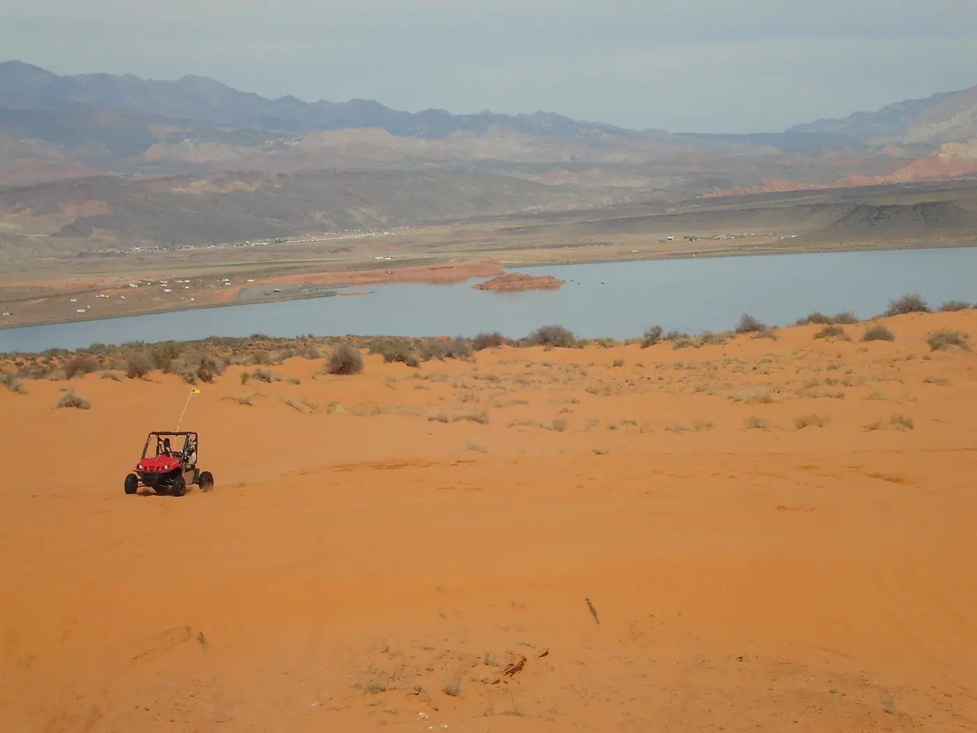 A red off-road vehicle on orange sand dunes above Sand Hollow Reservoir with mountains beyond