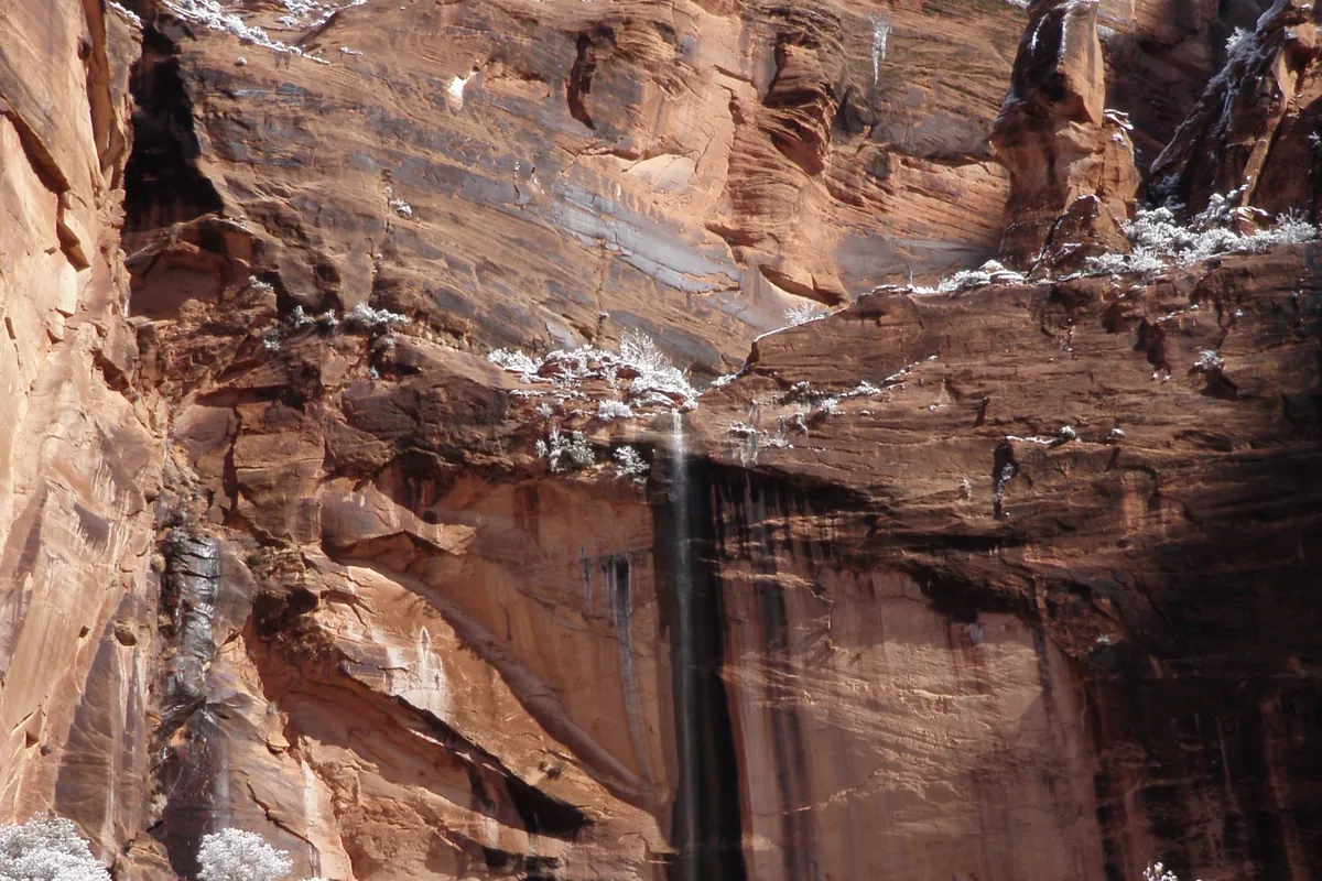 Thin waterfall running down a red sandstone wall near Weeping Rock, with snow-covered trees below