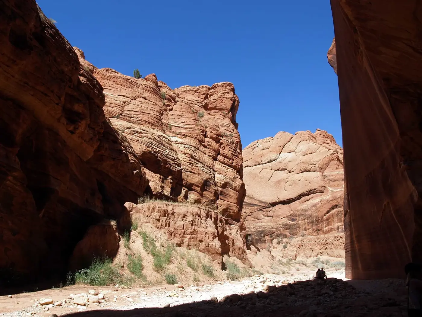 Hikers in shadow at the point where Wire Pass slot canyon meets Buckskin Gulch beneath tall red sandstone walls