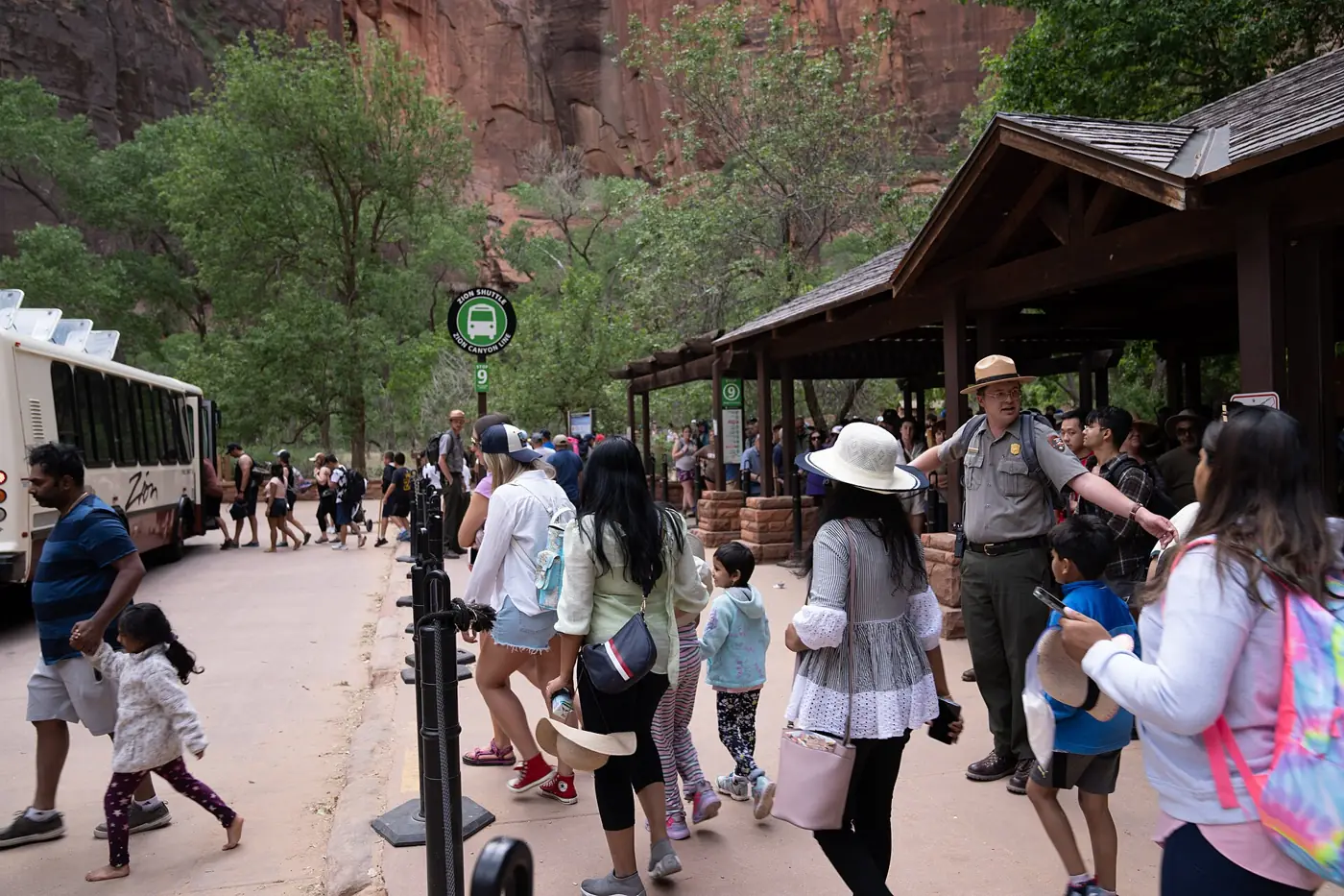 Visitors queuing beside a shuttle bus at a covered stop in Zion Canyon while a ranger directs the line
